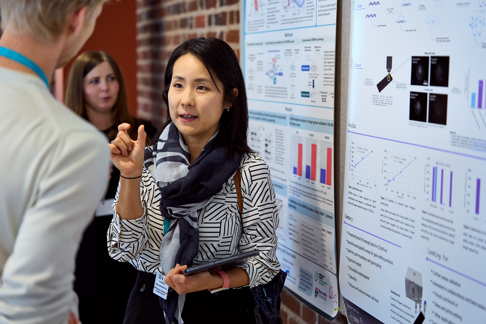 Two conference delegates discussing a scientific poster attached to a board that's suspended on a red brick background. 
