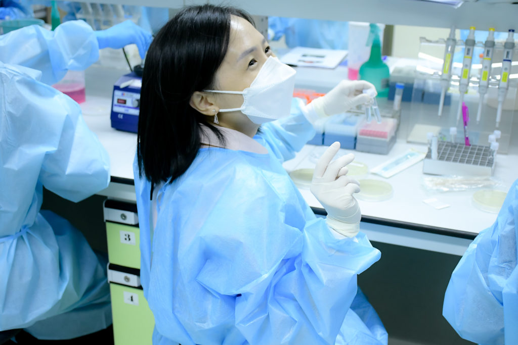 A colour photo of Polly Yap in a laboratory at Mahidol University. Polly is wearing a lab coat and gloves, seated at a bench, and holding two samples in one hand. She is looking out towards the left of the photo. Photo credit: Mahidol University