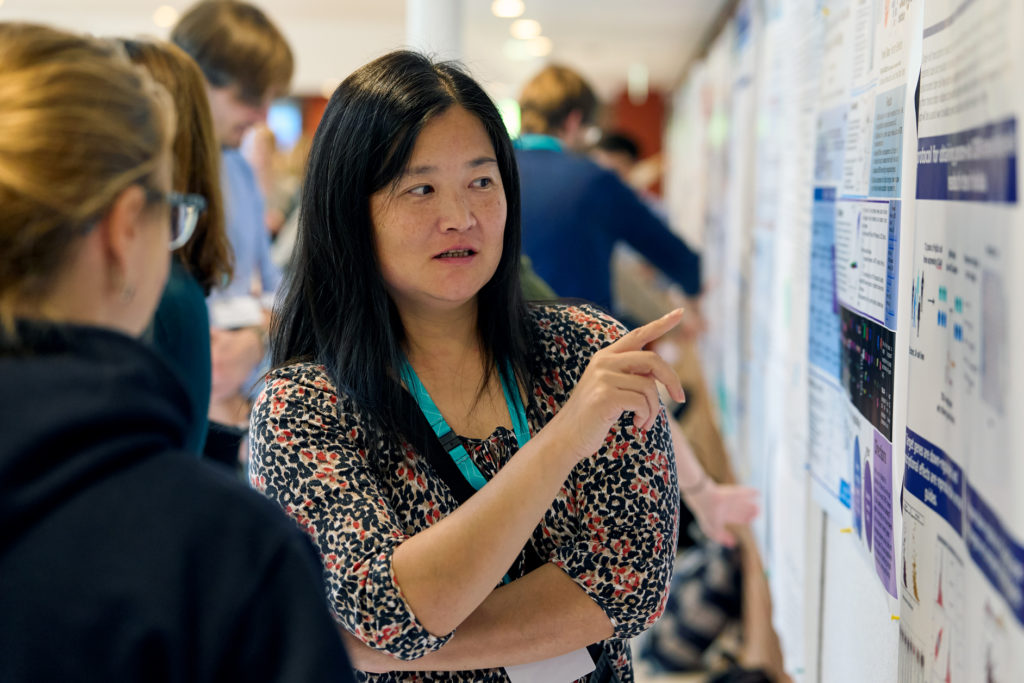 A colour photo of two participants discussing a scientific poster. One participant is pointing towards the poster, presenting her research insights to the other participant. 