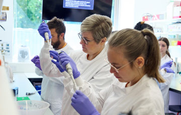 Three wet lab course participants taking part in a practical laboratory session. Each participant is standing at the bench and holding a pipette.