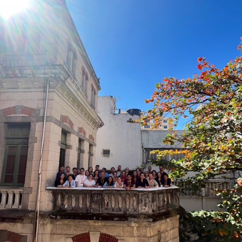 Single Cell Genomics course participants standing on a stone balcony of a former residence of Oswaldo Cruz, a pioneering Brazillian physician and epidemiologist. There is a tree in view, and the sun is shining over the building. 