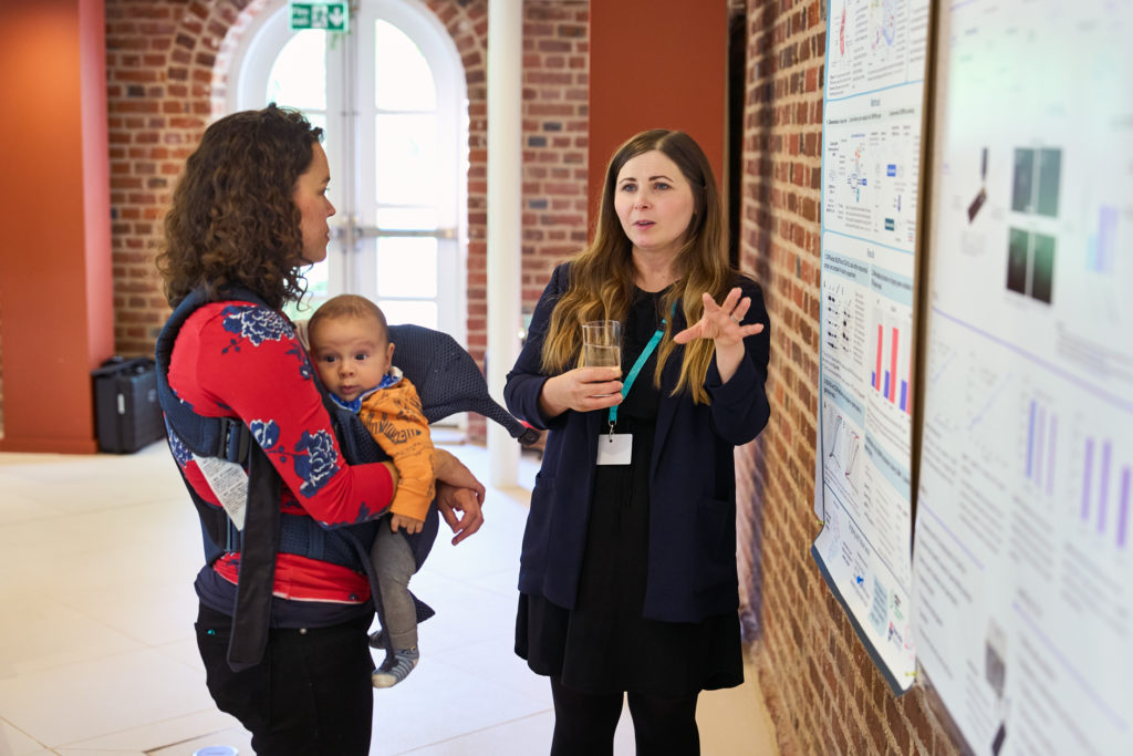 Two conference delegates chatting during a poster session. One delegate is holding a baby, the other delegate is pointing towards her work on a poster board.