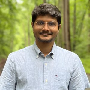 Dr Sounak Sahu facing forwards and smiling. He is wearing glasses and a light blue shirt. He is standing in front of trees. 