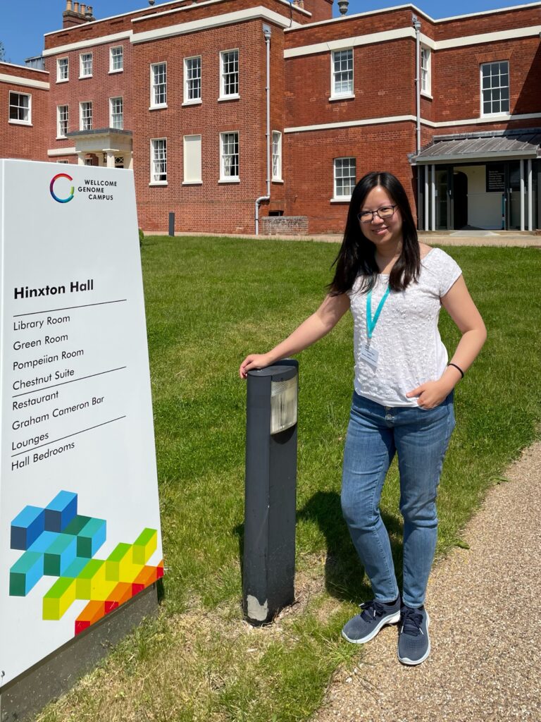 Dr Khánh-Dung ‘KD’ Nguyễn standing outside in front of Hinxton Hall. She is leaning on a post and smiling. 