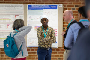 Delegates attending the 2022 Microbiome Interactions in Health and Disease poster session. One delegate is standing in front of his poster, pinned to a board. Three other delegates are looking at the poster. 
