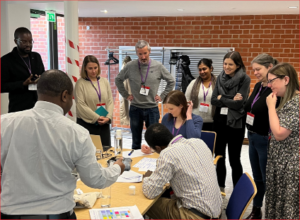 Participants working on a group exercise at our 2022 How to Teach Genomics workshop. Some are seated around a table with paperwork in front of them. Other participants are standing behind the seated delegates, observing what they writing.