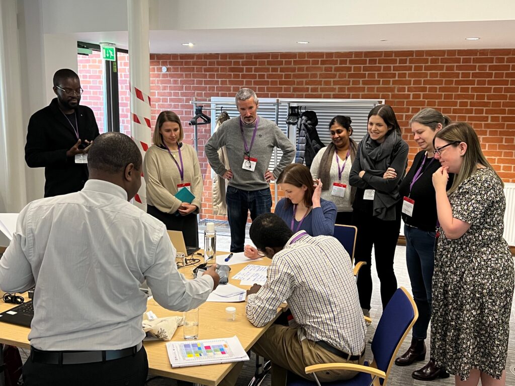 A group of T3connect trainees participating in a group exercise. They are gathered around a classroom table. There are pens, paper, laptops and water bottles on the table.  