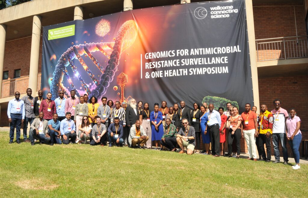 A group of participants standing underneath a banner promoting the Genomics for Antimicrobial Resistance Surveillance and One Health symposium. They are outside of a building, standing on grass. 