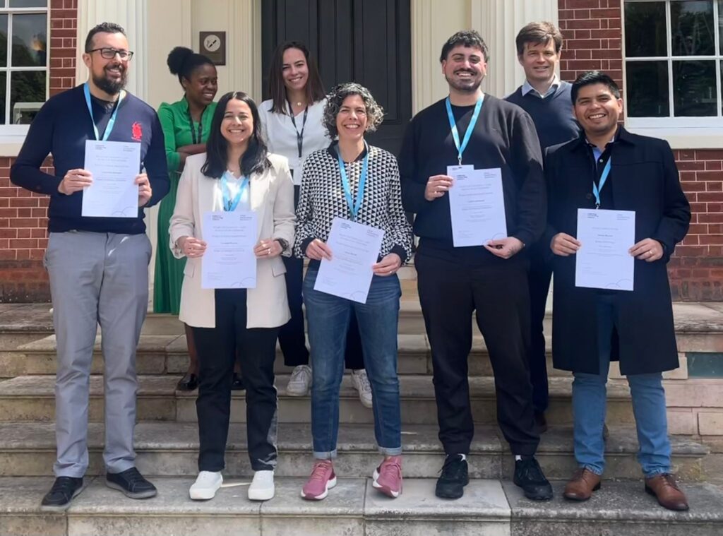 Grant writing champions standing on the steps of Hinxton Hall, a heritage building with red brickwork. Each of the five winners are holding up an award certificate. They are joined in the background by two single cell course organisers, and David Adams, a course trainer.