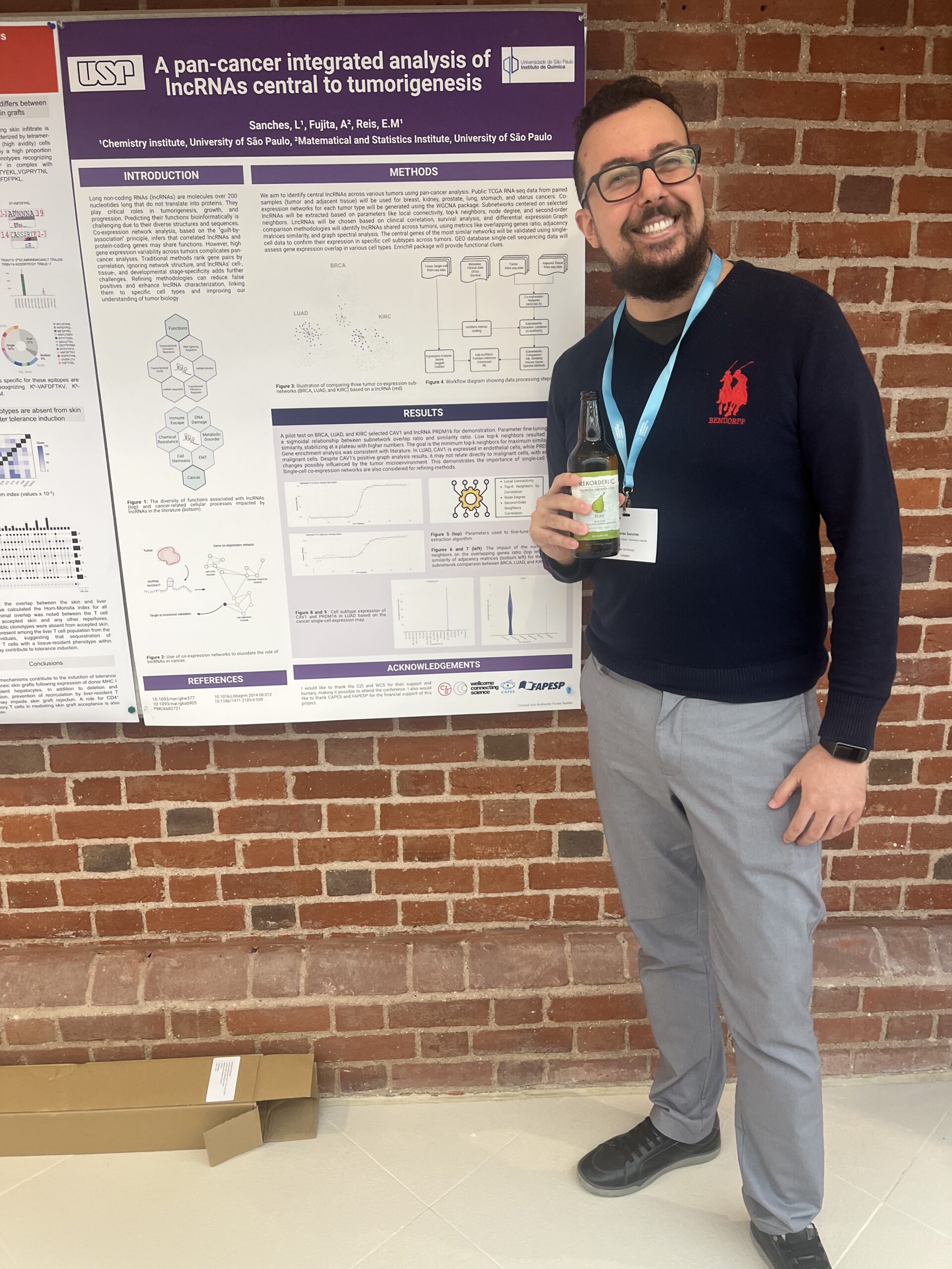 Leonardo Sanches standing in front of his poster at the conference. He is holding a drink in his right hand and smiling. 