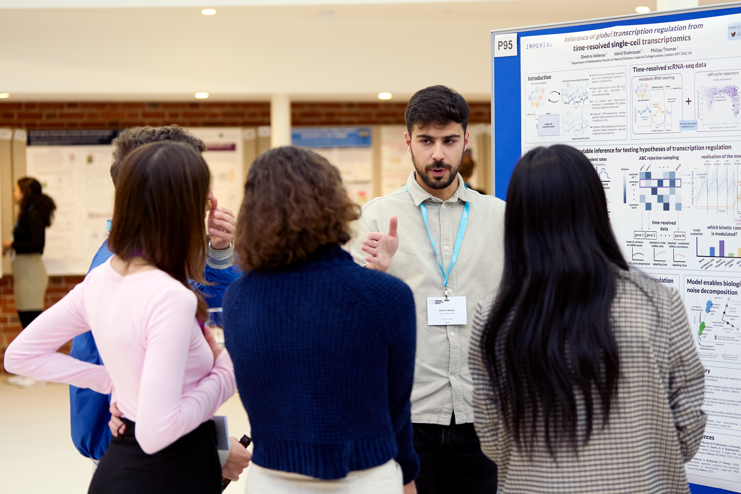 Four conference participants during a poster session. One is presenting a poster pinned to a blue board. The other three are listening. 