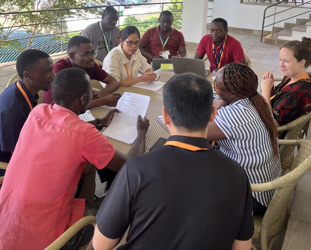 Participants seated at a table on an outside patio area in Kenya. They are collectively discussing their course work. 