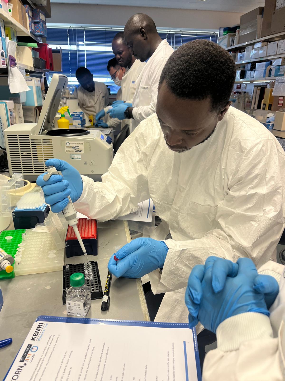 A course participant in a training laboratory engaged in a pipette exercise. They are wearing a white lab coat and blue safety gloves.