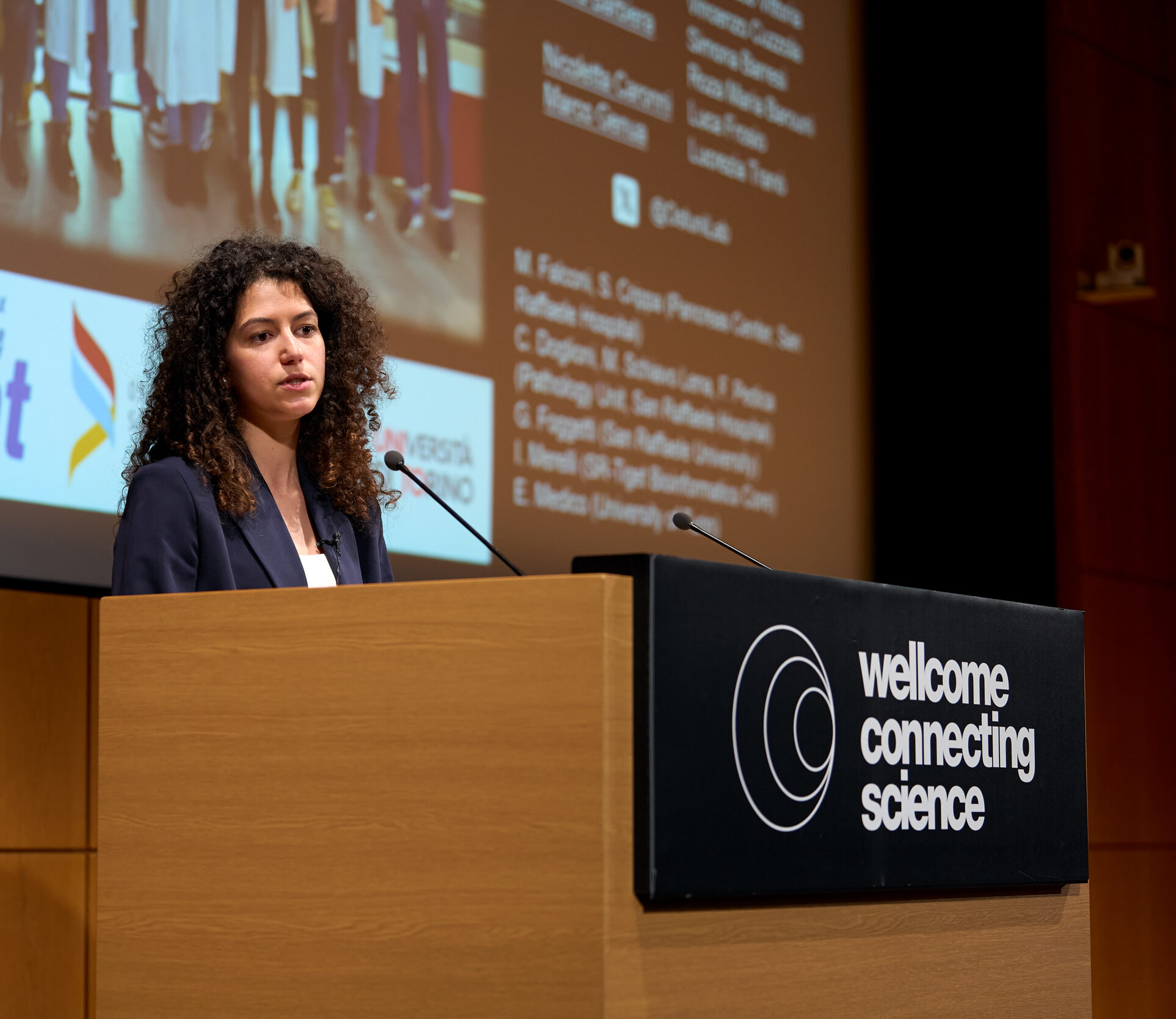 Conference speaker standing behind a branded podium on an auditorium stage. The speaker has a set of slides displayed on a screen in the background. The podium has the Wellcome Connecting Science logo printed on the front.
