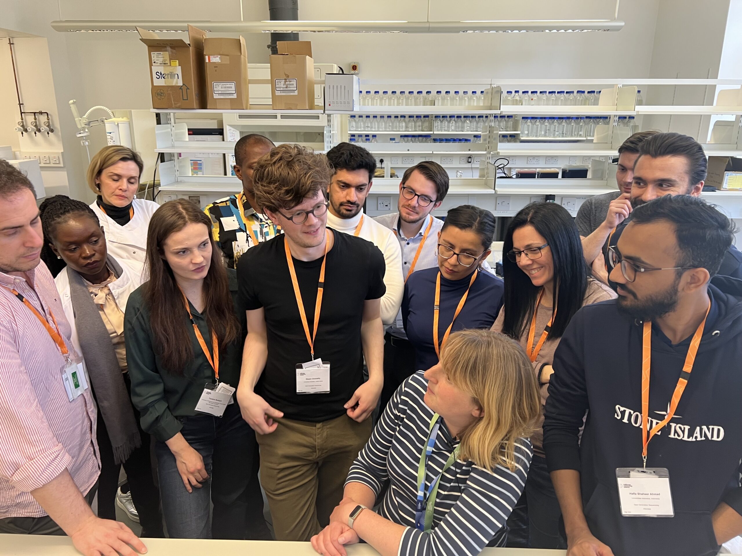 Course participants and instructors standing as a group in the training lab. There are racks holding lab equipment in the background. One of the instructors is leaning on a bench surface at the front of the photo.