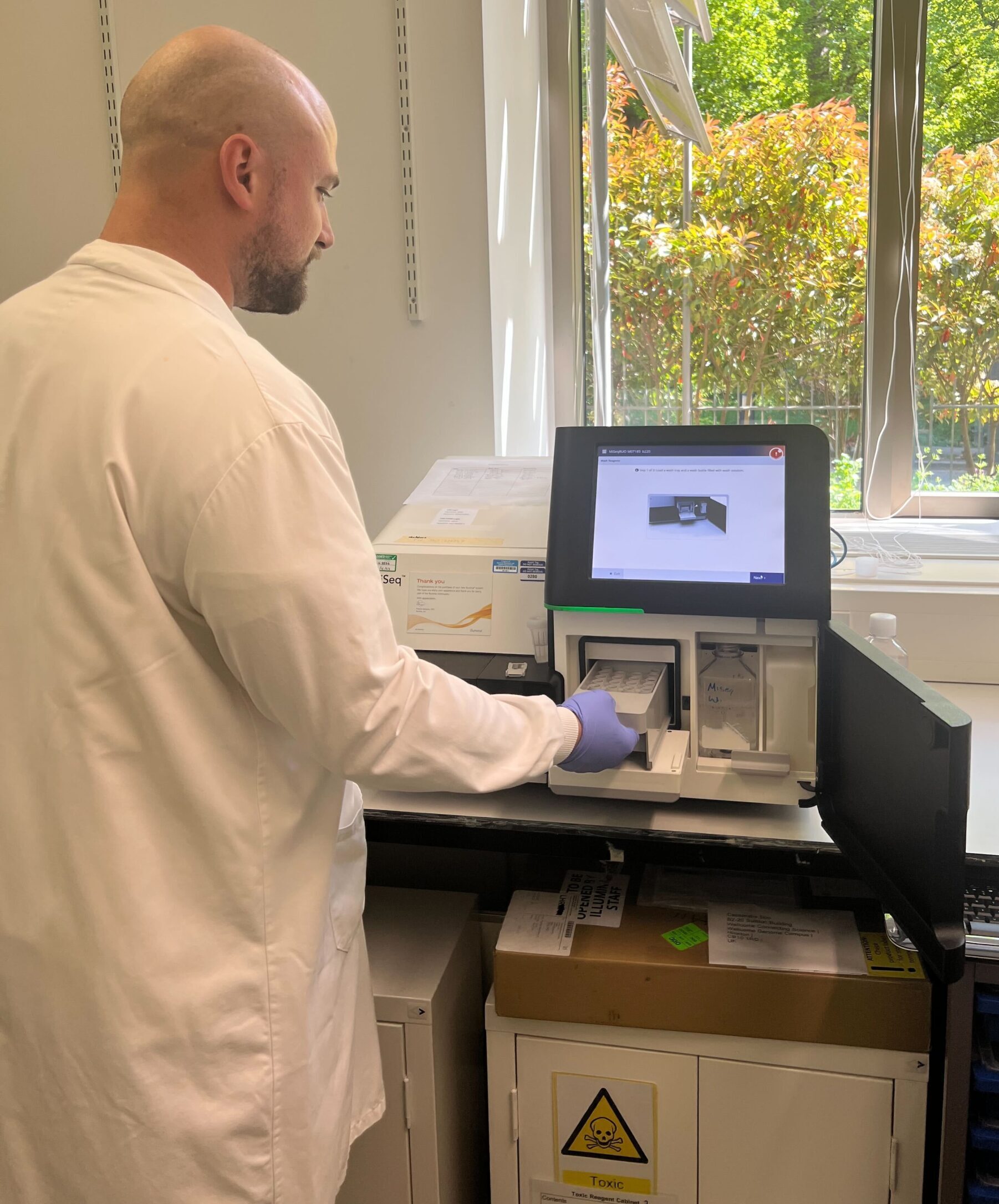 Aaron Dean, Laboratory Technical Officer, preparing our Illumina MiSeq sequencer for short-read sequencing. He is wearing a white lab coat and blue safety gloves.