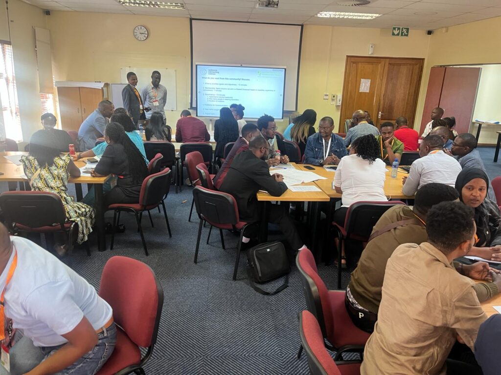 Wellcome Connecting Science Several groups of participants seated classroom style, at the 2024 AMR Symposium, South Africa. A lead presenter is standing at the front of the room, next to a projector.