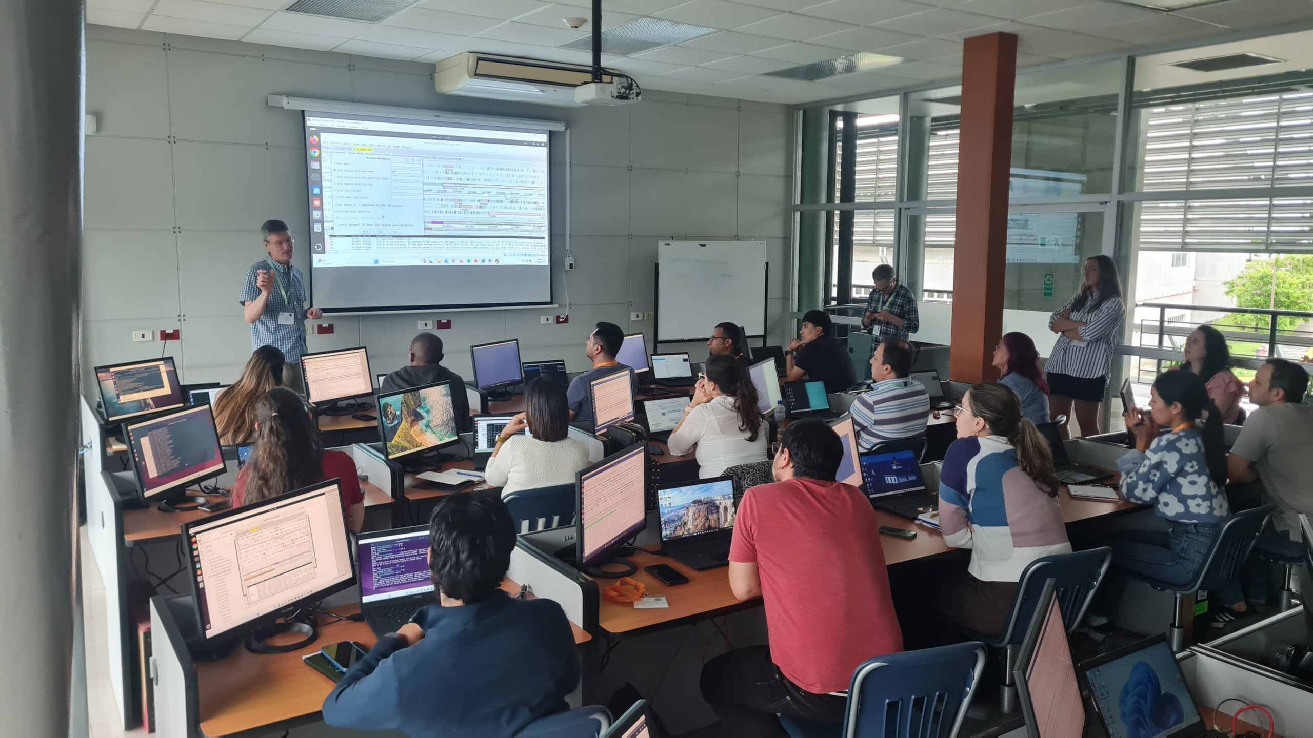 A diverse group of course participants attentively participating in a computer training session in a modern classroom with multiple monitors and a projected screen.