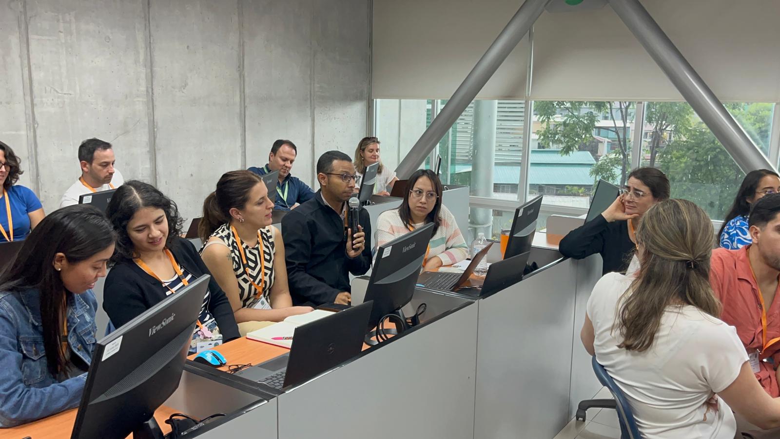 A diverse group of course participants engaged in a classroom-based training session, seated at desks with laptops and monitors, one person speaking into a microphone.