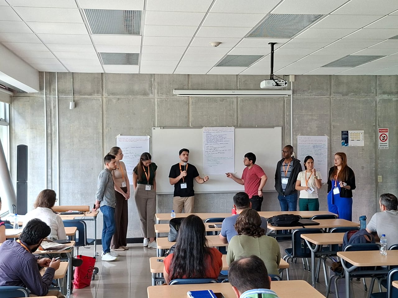 A diverse group of eight people standing and presenting in front of a classroom whiteboard with notes, while an audience sits at desks facing them.