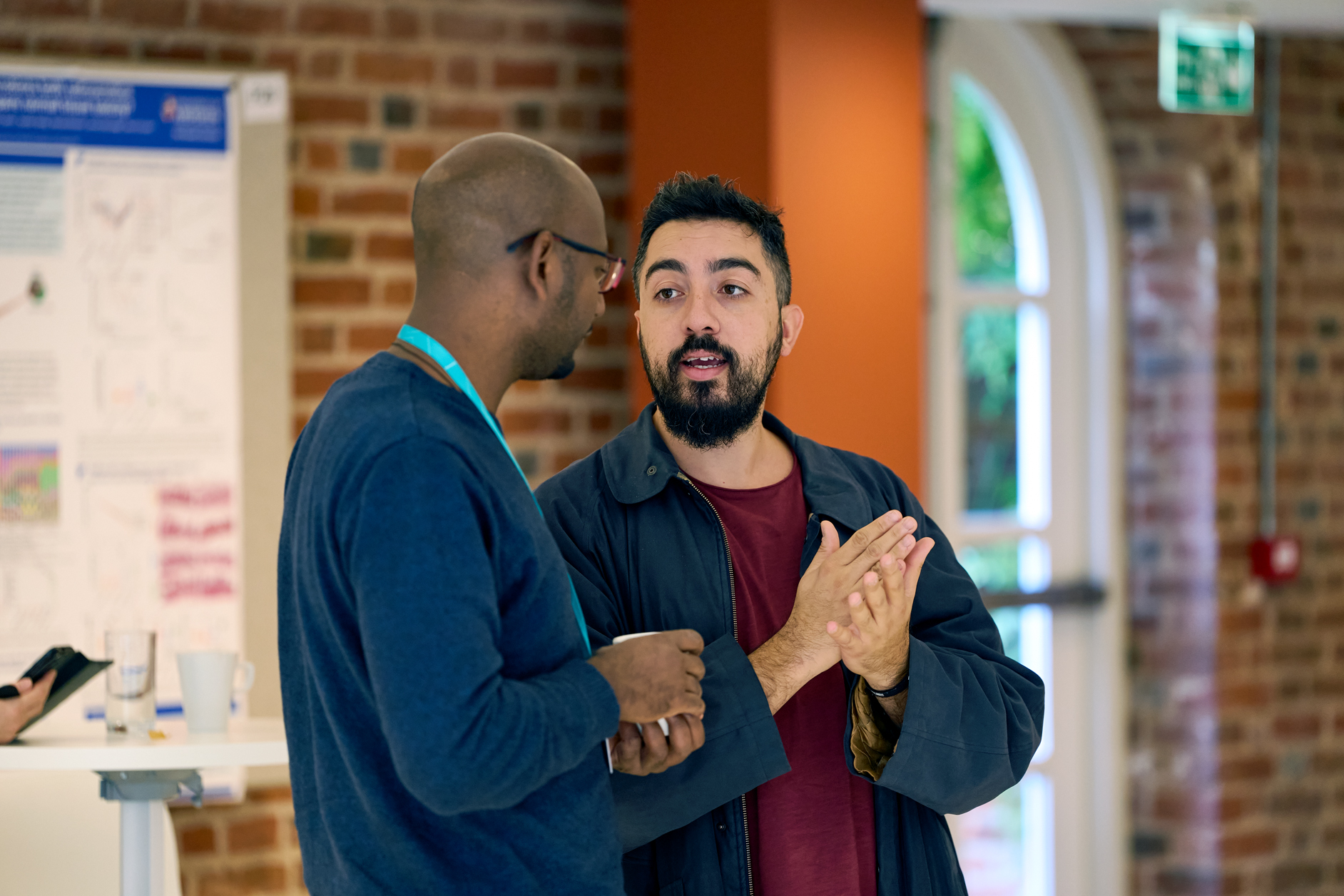 Two conference delegates engaged in conversation at a networking break, with a scientific poster pinned to a brick wall in the background.