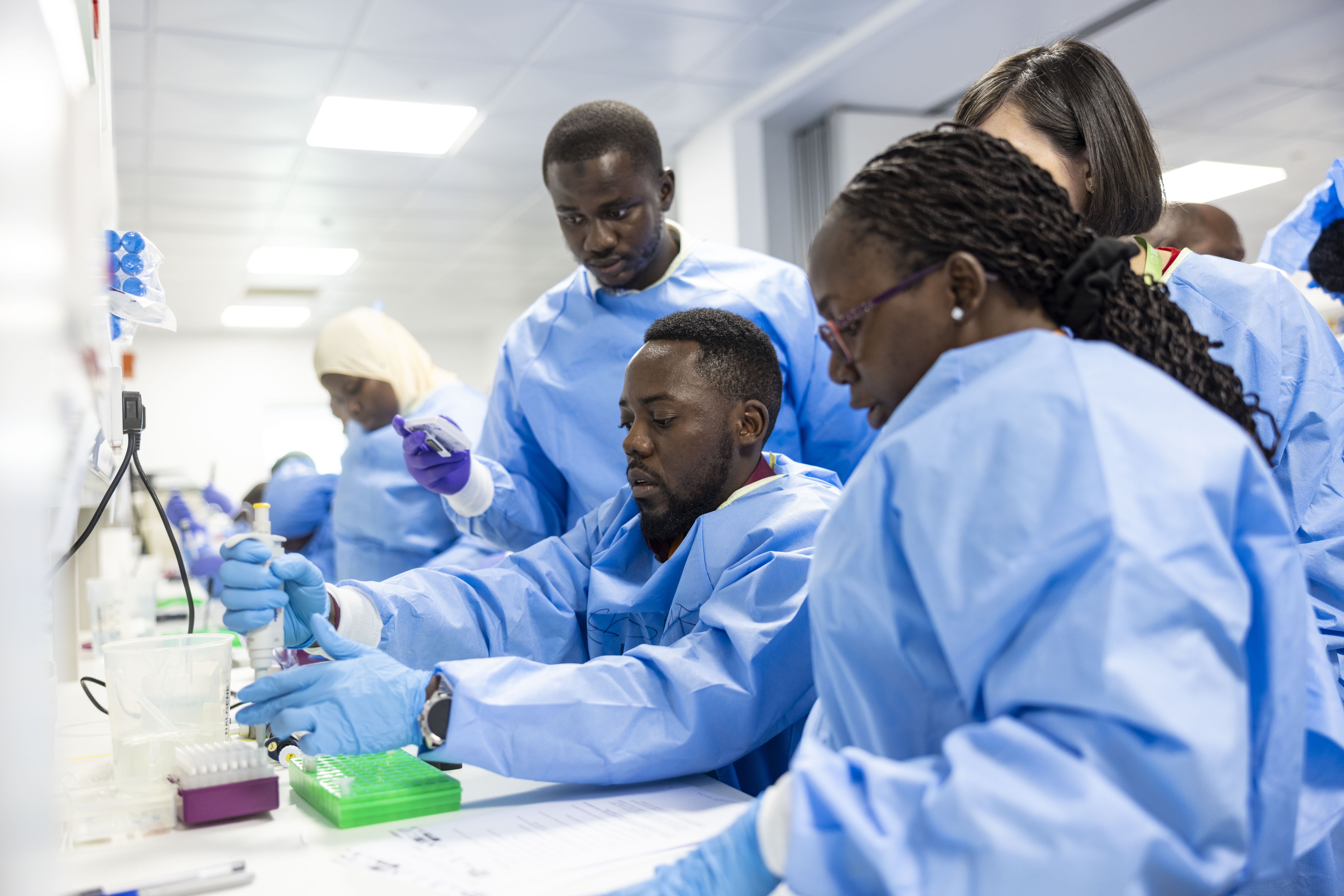 A group of course participants wearing blue lab coats and gloves collaborating in a laboratory setting, using pipettes during a training experiment.